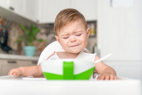 Síró kisgyerek egy asztalnál ül, előtte étel